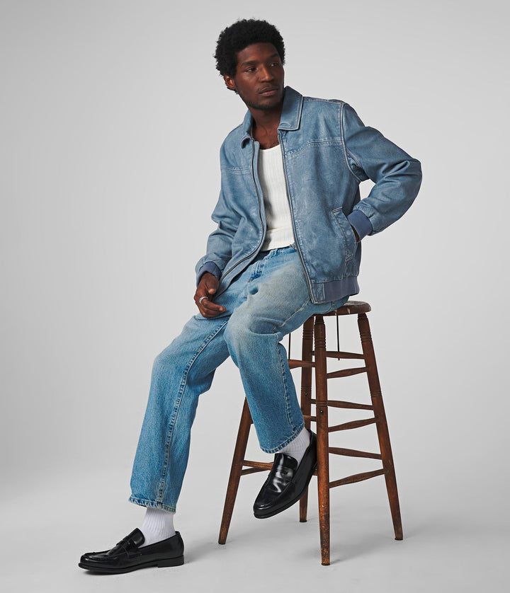 A man sitting, wearing a Nash Denim Leather Bomber with a zipper and two welt body pockets, paired with a white rib knit shirt.